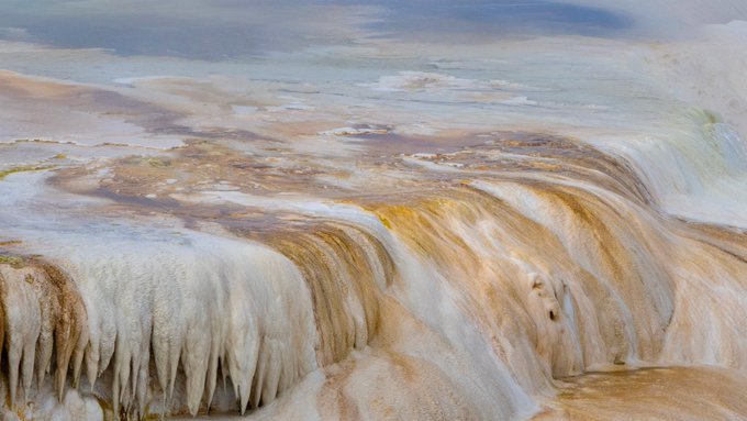 A close-up photograph of a terraced geological formation featuring white, cream, and rust-colored mineral deposits with cascading water flowing over the edges, resembling travertine terraces.