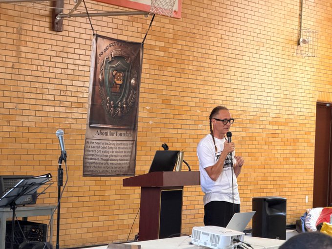 A man with glasses and a white t-shirt speaking into a microphone at a wooden podium in a brick-walled room, with an 'About Our Foundation' plaque hanging on the wall behind him.