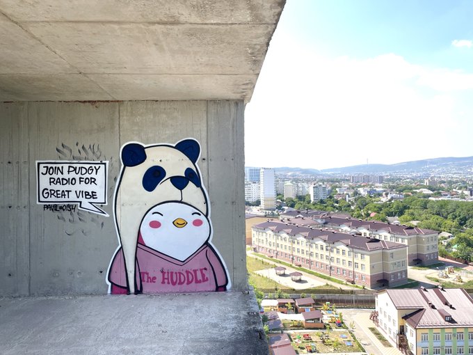 A street art mural of a penguin hugging a smaller penguin on a concrete wall reads "JOIN PUDGY RADIO FOR GREAT VIBE" and "The HUDDLE," overlooking a residential neighborhood, tying into the Pudgy Penguins NFT community referenced in the post.