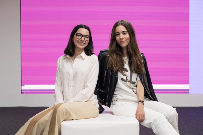 Two women posing together against a pink and white striped background, one wearing glasses with a white blouse and the other in a black leather jacket with white pants and a chain necklace.