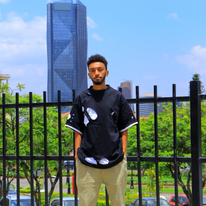 A young man poses confidently in front of a black metal fence with a modern skyscraper in the background, wearing a black oversized shirt with white graphics and khaki pants. The image complements his introduction as a 26-year-old content creator and Web3 community builder from Ethiopia, suggesting a professional yet approachable personal brand.