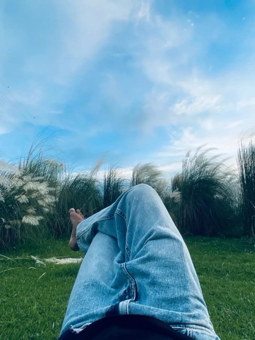 A first-person perspective photo of someone relaxing on grass wearing blue jeans, with their bare feet visible and tall grass/plants in the background under a blue sky with scattered clouds.