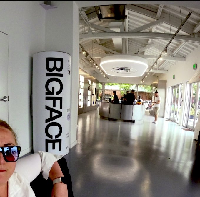 A selfie-style photo taken inside what appears to be the BIGFACE cafe, featuring a large cylindrical white sign with 'BIGFACE' printed vertically in black letters.