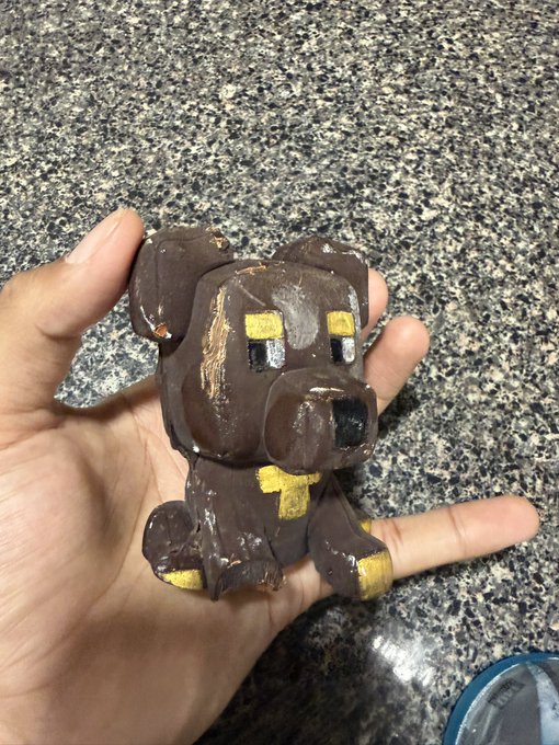 A hand holds a small, handmade clay figurine of a dog painted brown with yellow accents, showing its creation process on a speckled granite countertop.