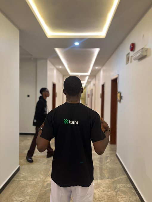 A person walking down a hotel hallway viewed from behind, wearing a black t-shirt with the 'lusha' logo on the back.