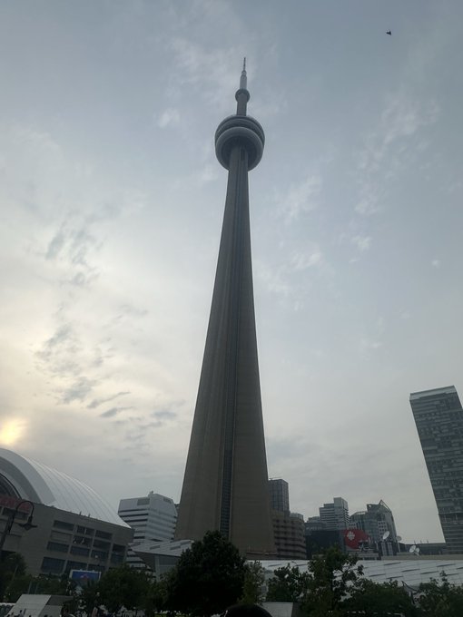 The image shows the CN Tower in Toronto under a cloudy sky, with a small drone visible to the right and a Canadian flag at its base. The caption cleverly ties the towering structure to the post's motivational message, using the tower as a visual metaphor for ambitious goals—both literally "going over your head" and serving as a soaring landmark that, much like the Finance Show's assistance, helps attendees reach new heights they couldn't achieve alone.