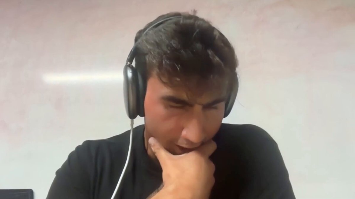 The image shows a man wearing headphones with his chin resting on his hand while looking down at something. The background appears to be a white wall or board. There are no texts present, so we can only see the man's facial expression and body language in the photo.