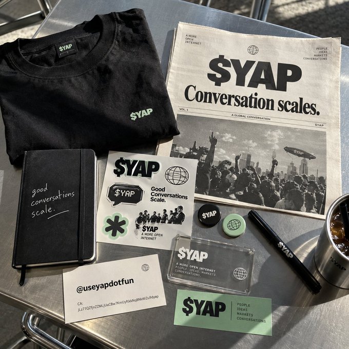 $YAP branded merchandise laid out on a metal table, including a black t-shirt, notebook reading 'good conversations scale.', stickers, pins, a pen, a newspaper-style flyer stating '$YAP Conversation scales.', and a handle tag '@useyapdotfun'.