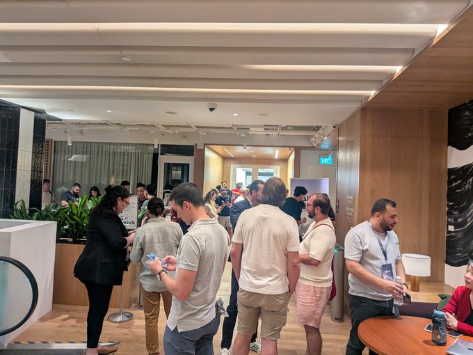 Indoor photograph of a crowded networking event or reception in a modern venue with wood-paneled ceilings, where numerous people in business casual attire are standing, mingling, and checking their phones near a bar area.