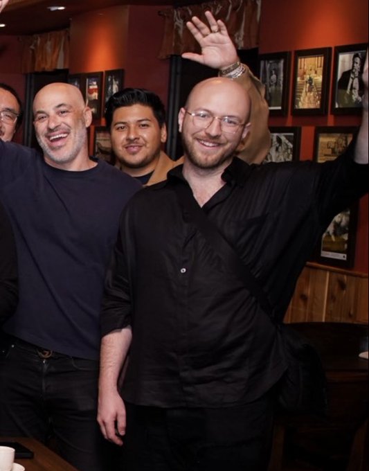 Three men smiling and posing indoors with one raising his hand, in what appears to be a restaurant with framed photos on the wall.
