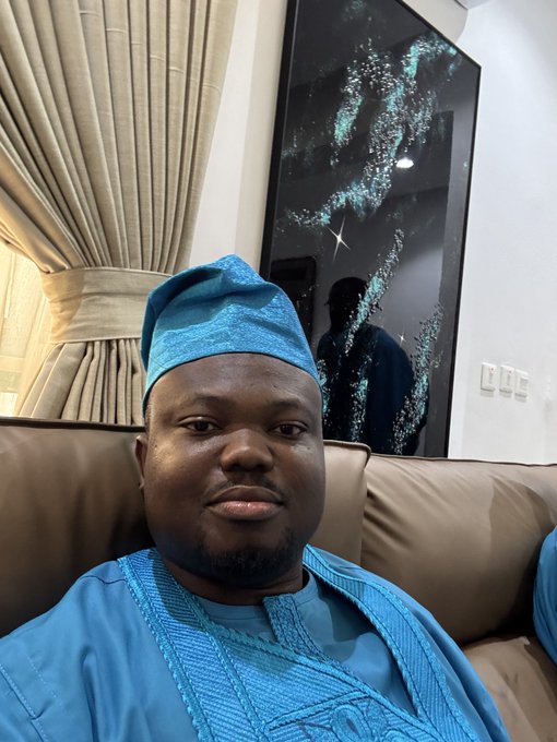 A selfie of a man wearing a blue embroidered traditional cap and matching blue shirt, sitting on a beige couch in front of a framed starry night artwork.