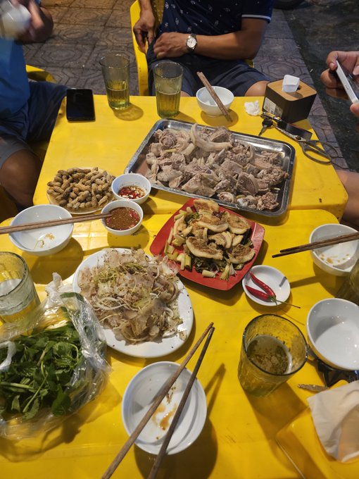 A yellow table set with Vietnamese food including a metal tray of boiled meat, spring rolls, fresh herbs, peanuts, dipping sauces, rice paper wrappers, chopsticks, and glasses of tea.