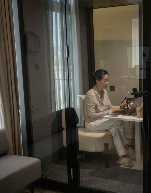 A woman in loungewear sits inside a glass-walled private booth or pod, working on a laptop at a small table in a dimly lit room.