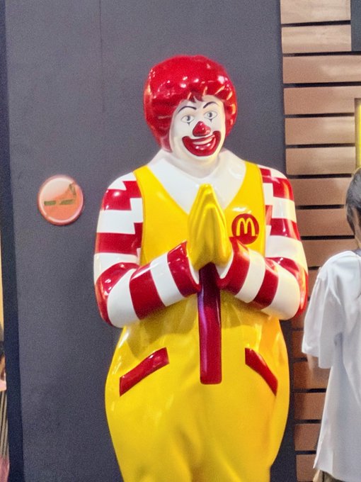 A Ronald McDonald statue in McDonald's signature yellow and red outfit standing with hands clasped in prayer position, positioned against a dark wall with a person in a white shirt partially visible on the right.