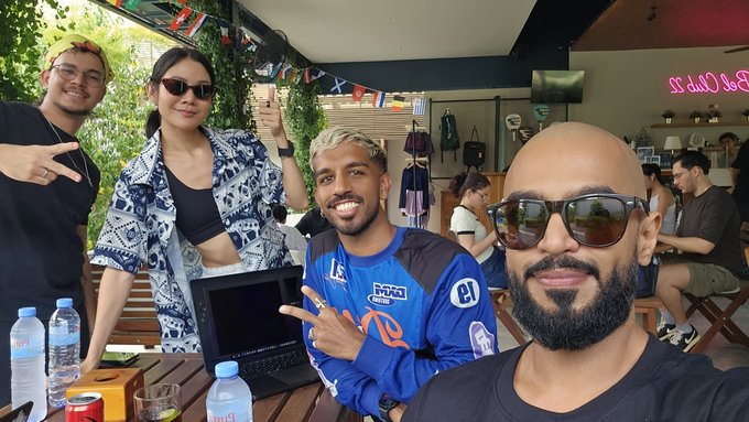 Four people posing together at an outdoor cafe with international flags overhead, including a woman in a blue patterned shirt, a smiling man in a blue jersey, and a bearded man in sunglasses taking a selfie.