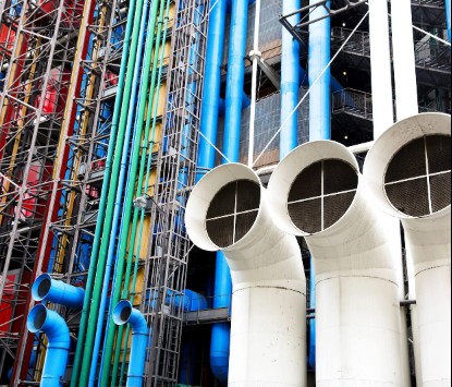 An industrial exterior view shows large white ventilation pipes alongside colorful red, green, and blue pipes running vertically against a building facade.