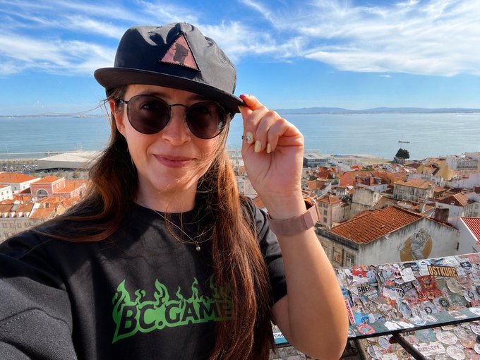 A woman wearing sunglasses and a cap takes a selfie overlooking a coastal city, wearing a BC.GAME branded t-shirt.