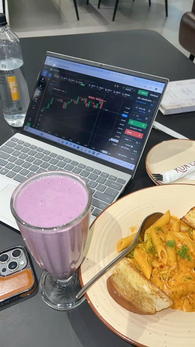 A lifestyle photo showing a workspace with a laptop displaying a trading chart, a pink smoothie in a glass, a bottle of water, and a plate of penne pasta with bread, with a phone and AirPods visible nearby.