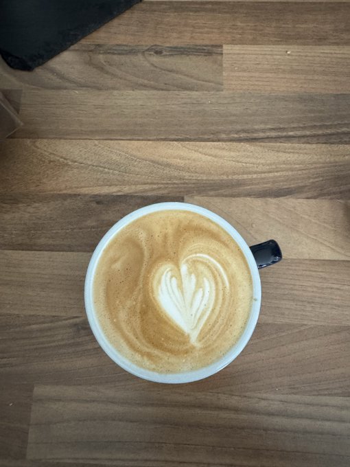 A freshly made cappuccino with beautiful latte art in a heart pattern sits on a wooden table, perfectly matching the "Coffee first!" sentiment of the morning motivation post.