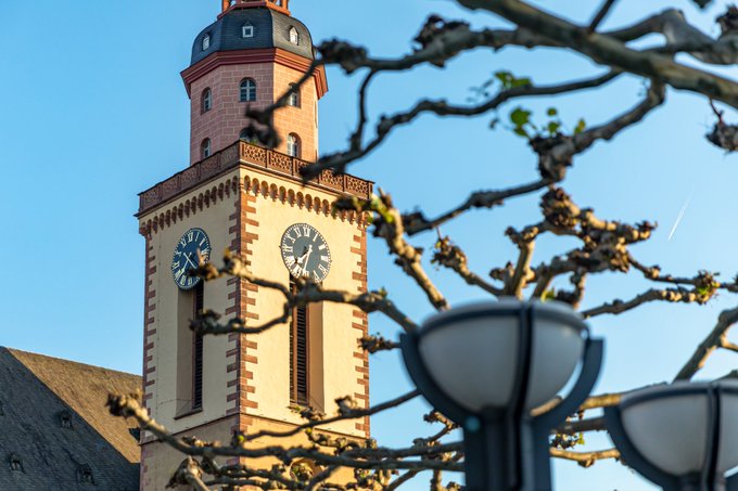 A photograph of a tall church or clock tower with two visible clock faces, framed by bare tree branches against a clear blue sky.