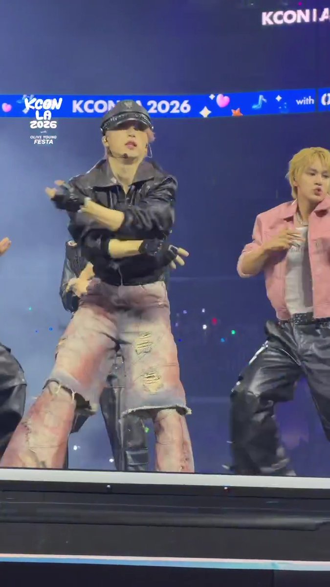 Two JO1 members perform energetically on stage at KCON LA 2026, with the "KCON LA 2026 with OLIVE YOUNG FESTA" banner visible behind them, capturing the excitement of the COUNTDOWN STAGE on Day 3 at Crypto.com Arena.
