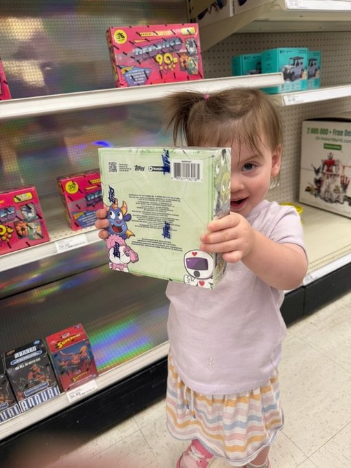 A young girl in a store holding up a Tokidoki Toki Moki Blind Box package in front of her face, with other toys visible on the store shelves behind her.