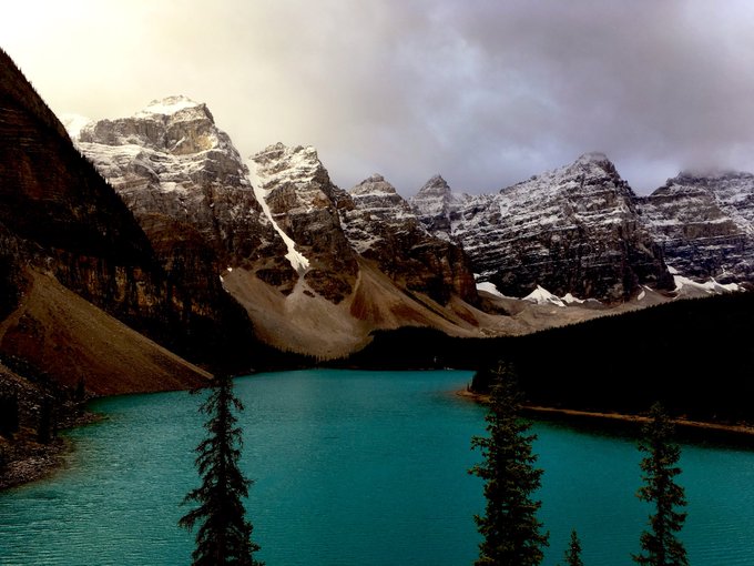 A stunning mountain landscape features snow-capped peaks, including a prominent pyramid-shaped mountain, rising above a vibrant turquoise lake with evergreen trees in the foreground under a cloudy sky.