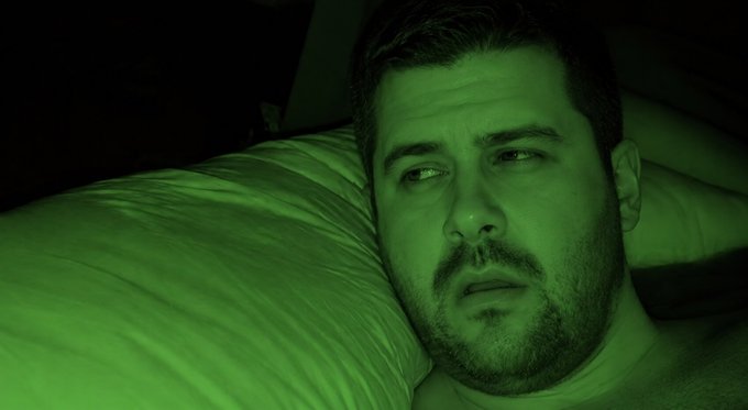 A close-up selfie shows a bearded man lying in bed, illuminated by a green night-vision-like light.