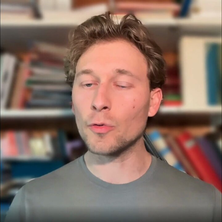 The image features a man wearing a gray shirt, standing in front of a bookshelf filled with numerous books. The man appears to be talking into a microphone or recording device, possibly discussing the importance of infrastructure for tokenized assets. The background is blurred, but there are several books visible on the shelf behind him.