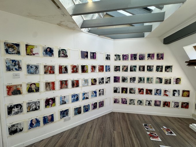 An art gallery installation displaying numerous small portrait artworks hanging on white walls beneath skylights.