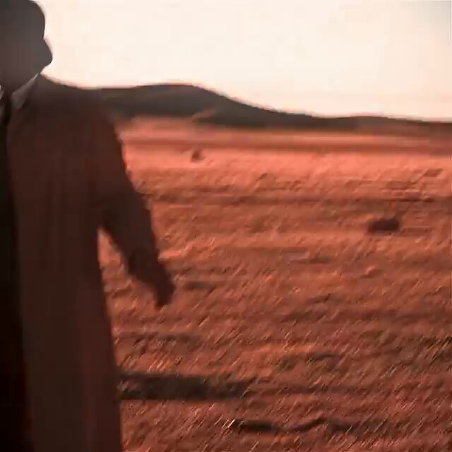 The image features a man standing in an open field with a sandy-colored landscape surrounding him. He appears slightly blurred, suggesting movement or distance between him and the camera angle. The scene takes place under a sunset, creating a beautiful, warm atmosphere that adds depth to the photograph.