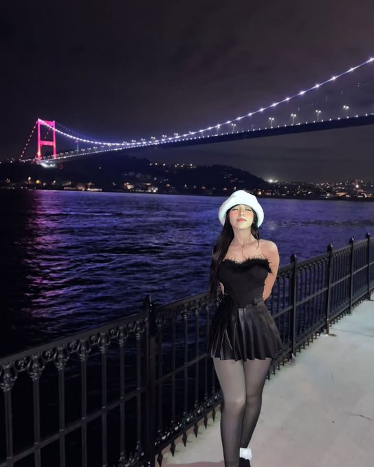A night-time portrait of a woman wearing a white fuzzy hat, black off-shoulder top, black pleated skirt, and sheer black tights, standing on a walkway with an illuminated suspension bridge in the background.