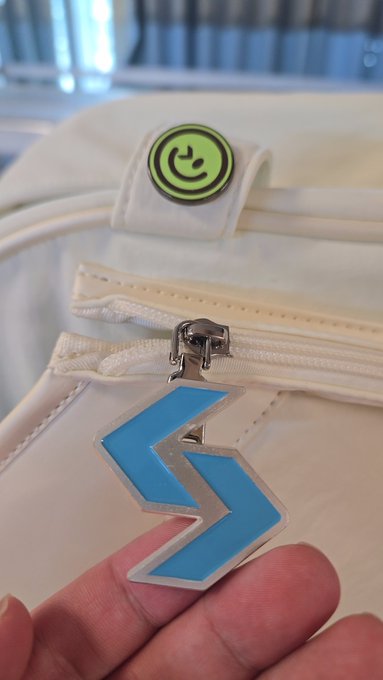 A close-up of a hand holding a blue chevron/arrow-shaped zipper charm attached to a cream-colored bag, with a small green smiley face tag visible above.