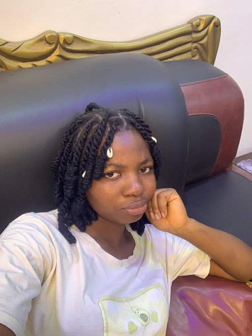 A young woman with braided hair and cowrie shell accessories takes a selfie while resting her head against a large black and maroon leather chair with an ornate gold headboard behind it.