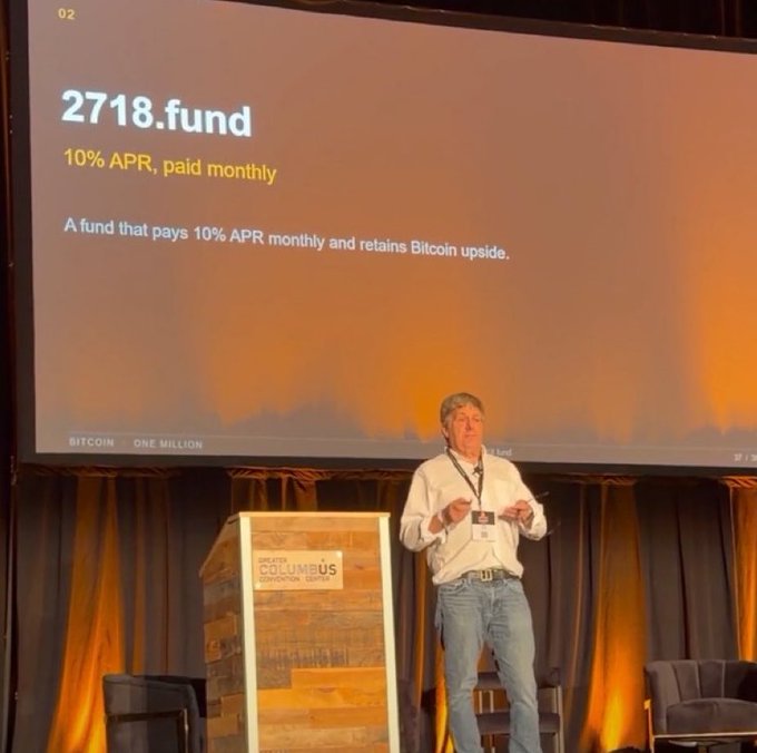 A man in a white shirt and jeans gives a presentation on stage at a 'Greater Columbus Convention Center' event, with a slide displaying '2718.fund - 10% APR, paid monthly - A fund that pays 10% APR monthly and retains Bitcoin upside.'