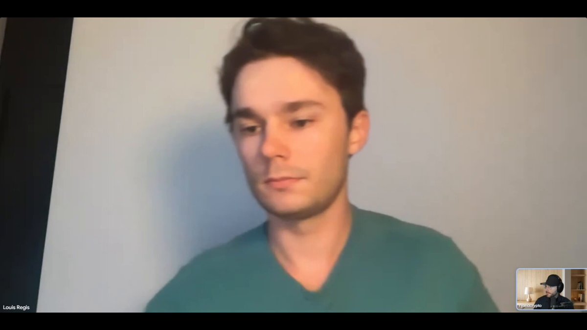 The image shows a man wearing a green shirt sitting at his computer, with a video call displayed on the screen. The man appears to be engaged in an interview or discussion about cryptocurrency trading. The background of the image features a white wall, which contrasts with the black frame around the window where the man is seated.