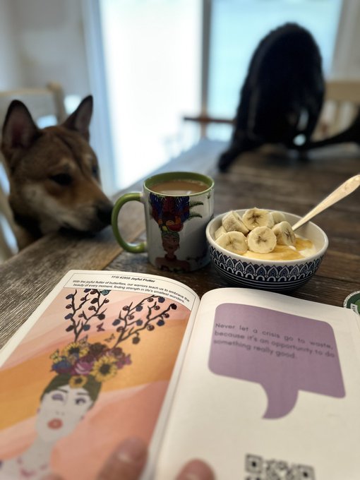 Cozy photo of a dog resting its head on a wooden table next to a coffee mug, a bowl of banana slices, and an open book, with a cat blurred in the background.