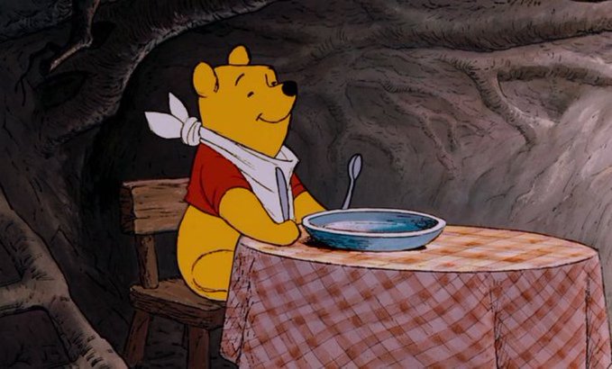 Winnie the Pooh sits at a small wooden table with a red checkered tablecloth, holding a spoon over a blue bowl of food inside a cave-like room.