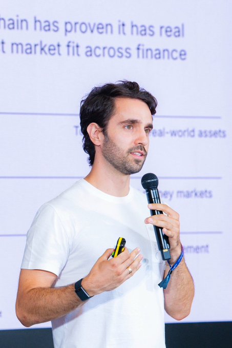 A man in a white t-shirt speaks into a microphone while presenting on stage, with text mentioning 'market fit across finance' visible behind him.