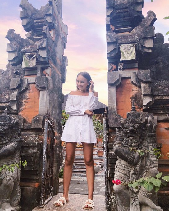 A young woman in a white off-shoulder dress standing between ornate Balinese stone gate pillars at sunset, with traditional guardian statues on either side.