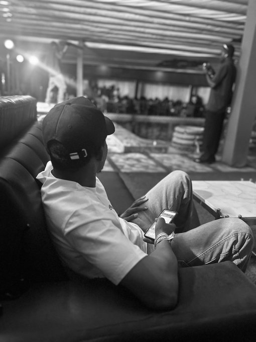 A black-and-white photo of a man wearing a cap and t-shirt lounging on a dark sofa while looking at a phone, with a large crowd visible in the background of what appears to be an event hall.