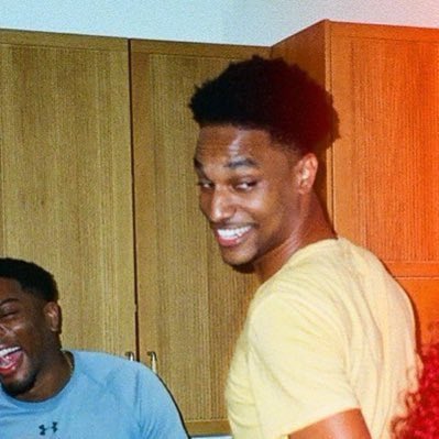 A photo showing a smiling young man with short dark hair wearing a yellow t-shirt, standing in a room with wooden cabinets, with another person laughing in the foreground.