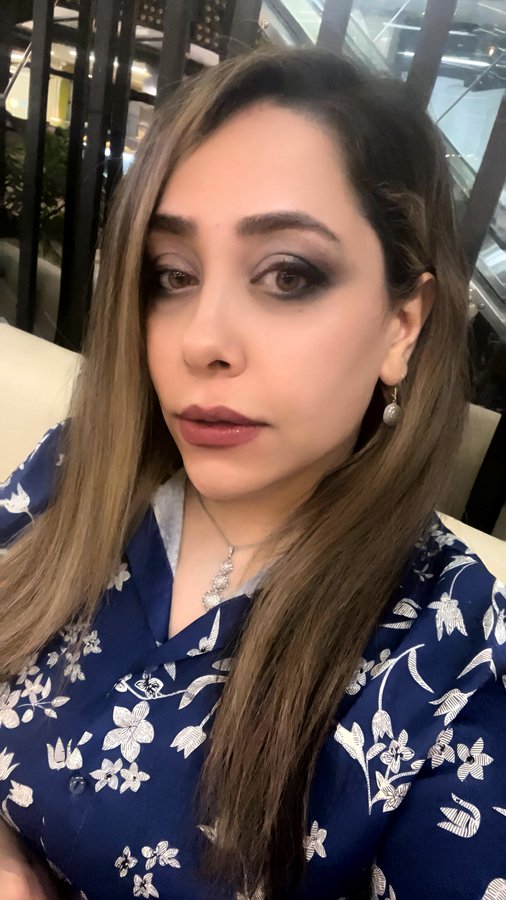 A woman with long brown hair and smoky eye makeup poses for a selfie, wearing a blue floral blouse and a silver pendant necklace in what appears to be a mall setting.