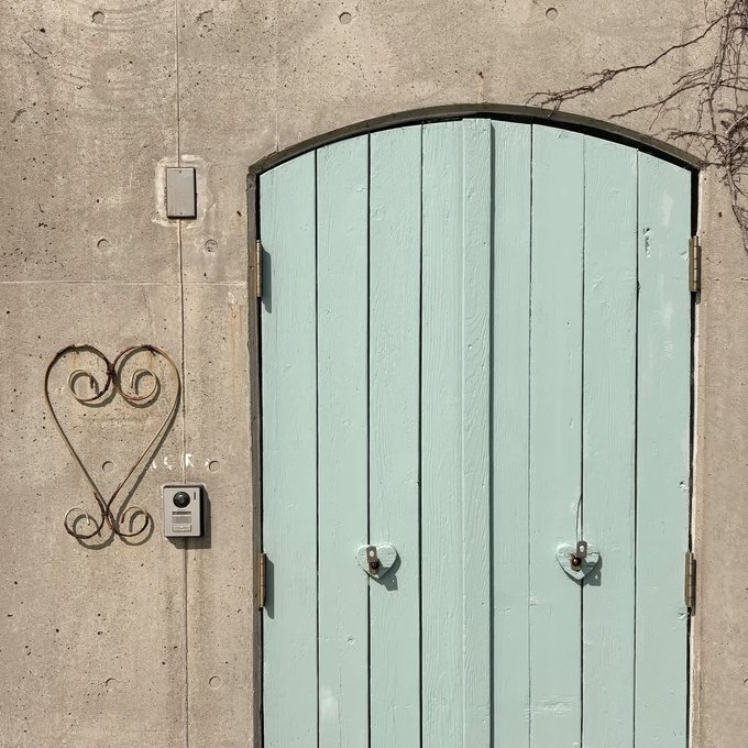 A pale mint-green wooden arched double door set in a concrete wall, featuring a decorative wrought iron heart-shaped ornament to its left.