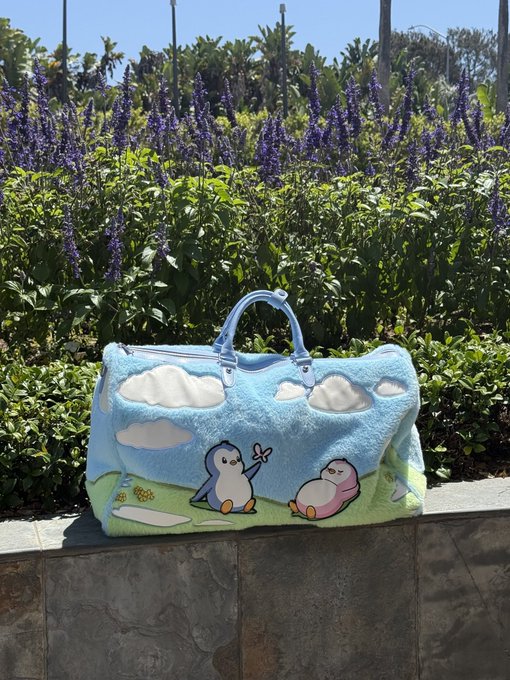 A light blue felt travel bag featuring a cute penguin character design, photographed outdoors against a backdrop of purple salvia flowers and greenery.