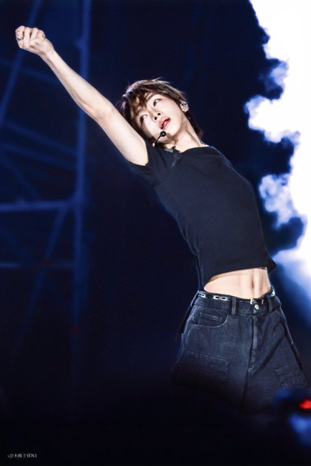 A performer with brown hair, wearing a black crop top and dark pants, poses dynamically on stage with one arm raised, illuminated by stage lights and pyrotechnics.