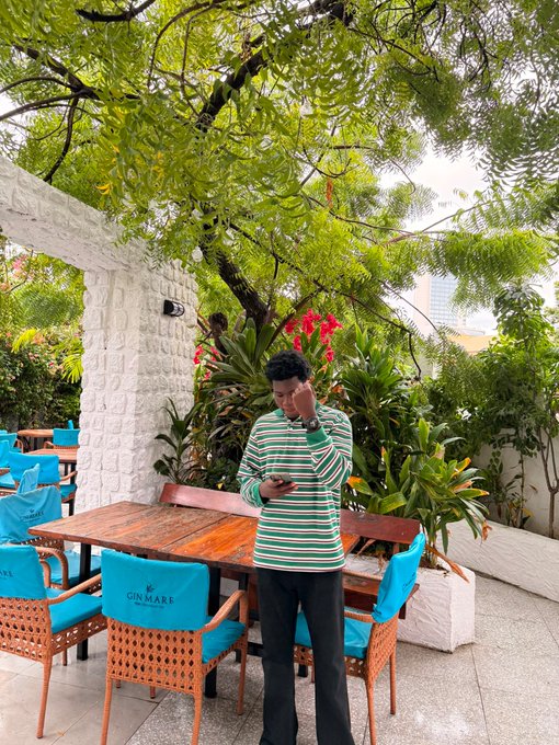 A young man wearing a green and white striped shirt stands at an outdoor patio table with blue "GIN MARA" chairs, looking down at his phone with one hand near his face, surrounded by lush green trees and plants.
