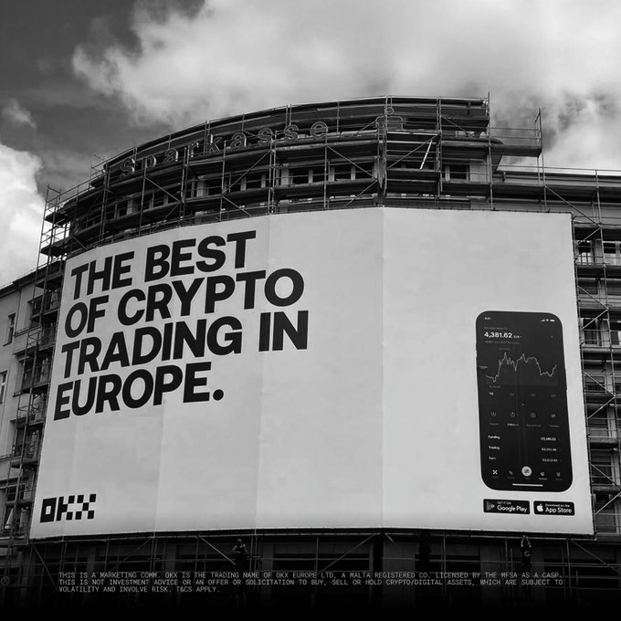 A black-and-white photo of a large billboard on a building (labeled 'Spirka.se') advertising OKX with the text 'THE BEST OF CRYPTO TRADING IN EUROPE.' alongside a smartphone showing the trading app and App Store/Google Play download badges.