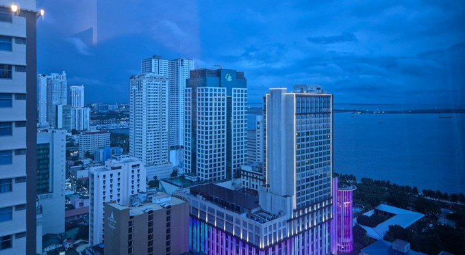 A dusk view of a coastal city skyline with modern high-rise buildings, some featuring purple lighting, set against a cloudy blue sky and a waterfront.