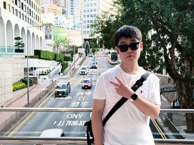 The image features a young man wearing sunglasses, standing next to a railing overlooking a busy city street filled with cars and buses. He has his hand raised in a peace sign gesture. The caption of the image includes an interesting statement about Binance, which is likely related to the person's interest or passion for cryptocurrency trading.